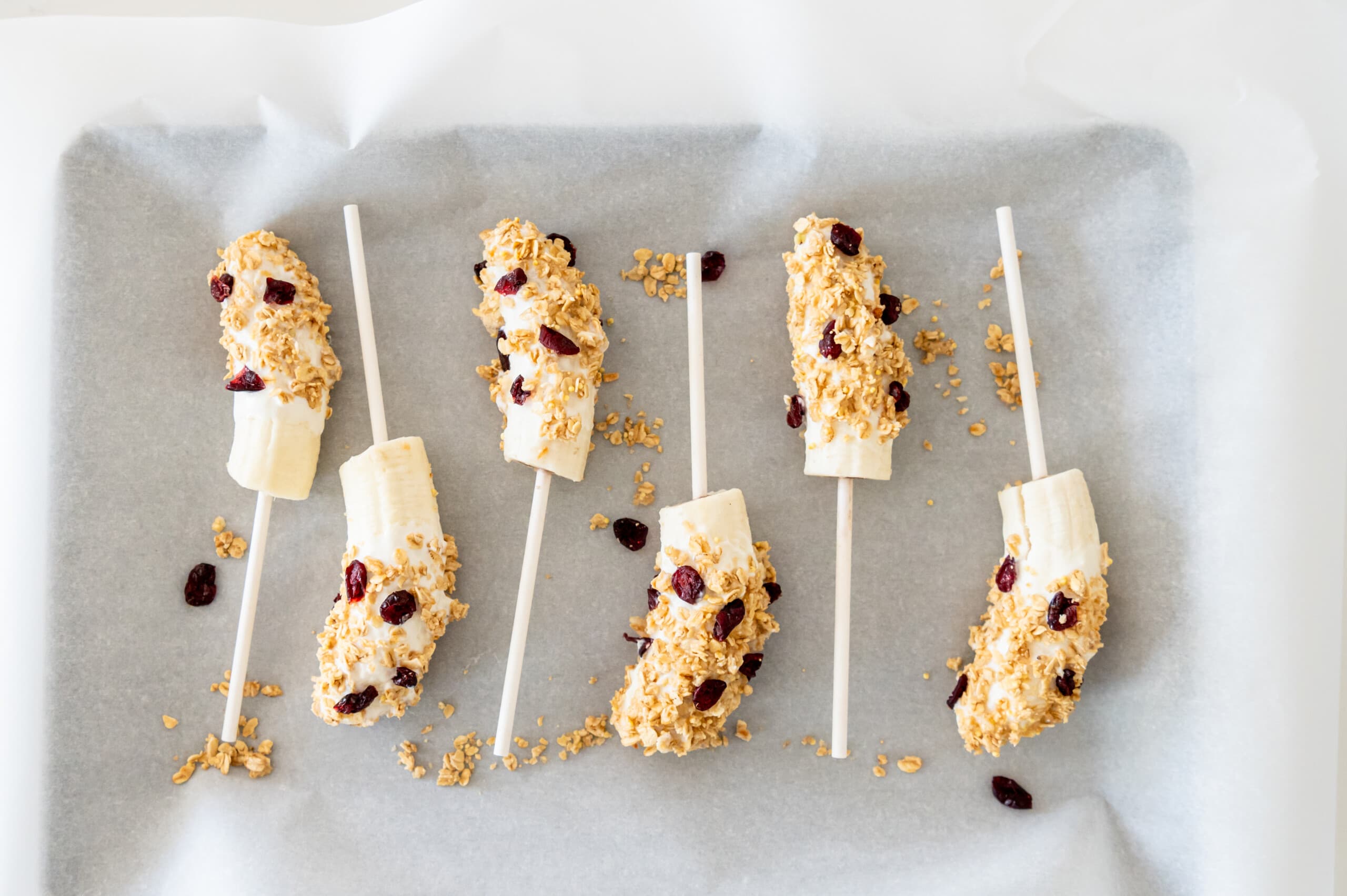 A parchment paper lined cookie sheet with yogurt covered banana pops with granola and dried cranberries