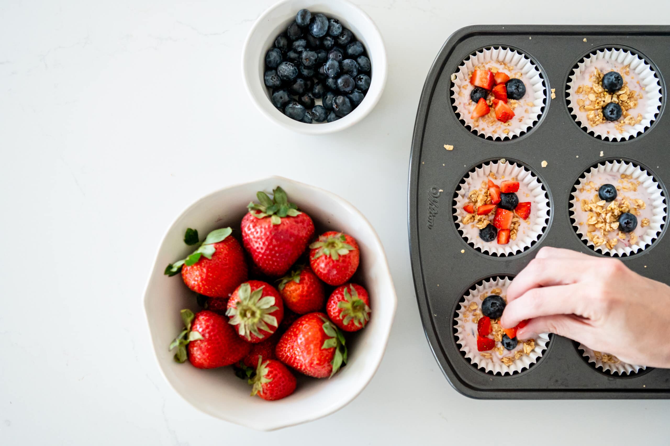 Hand sprinkling berries and granola on top of yogurt in muffin tins.