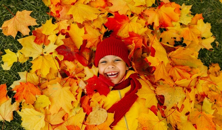 A girl with a wide smile lies on a carpet of red and yellow leaves in an autumn park.
