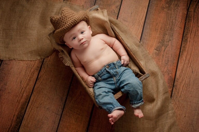 A four month old baby boy wearing a crocheted cowboy hat. He is lying in a wooden crate lined with burlap. Shot in the studio on a rustic, wood background.