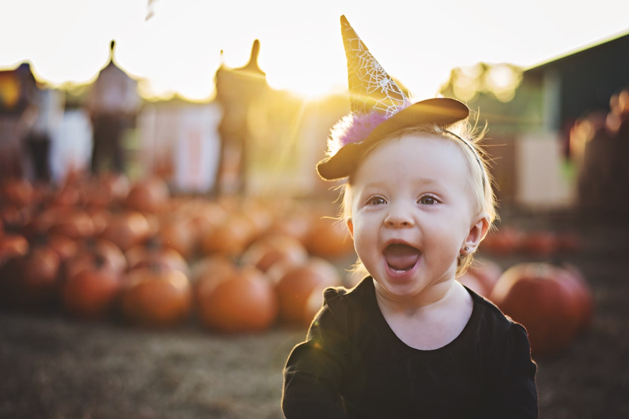 Baby girl dressed in purple and black and a witch hat on a headband crawls through a pumpkin patch on Halloween.