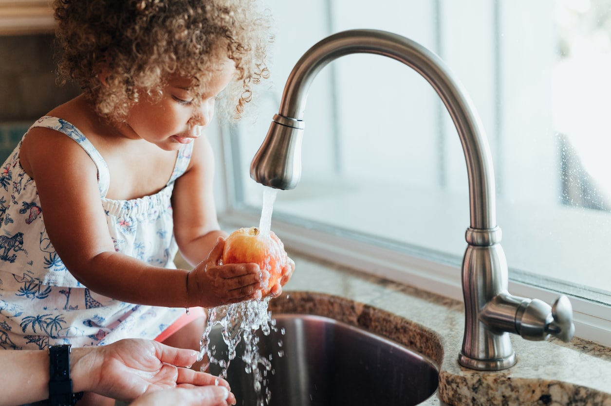 Cute toddler with mom together at home washing apples in the sink