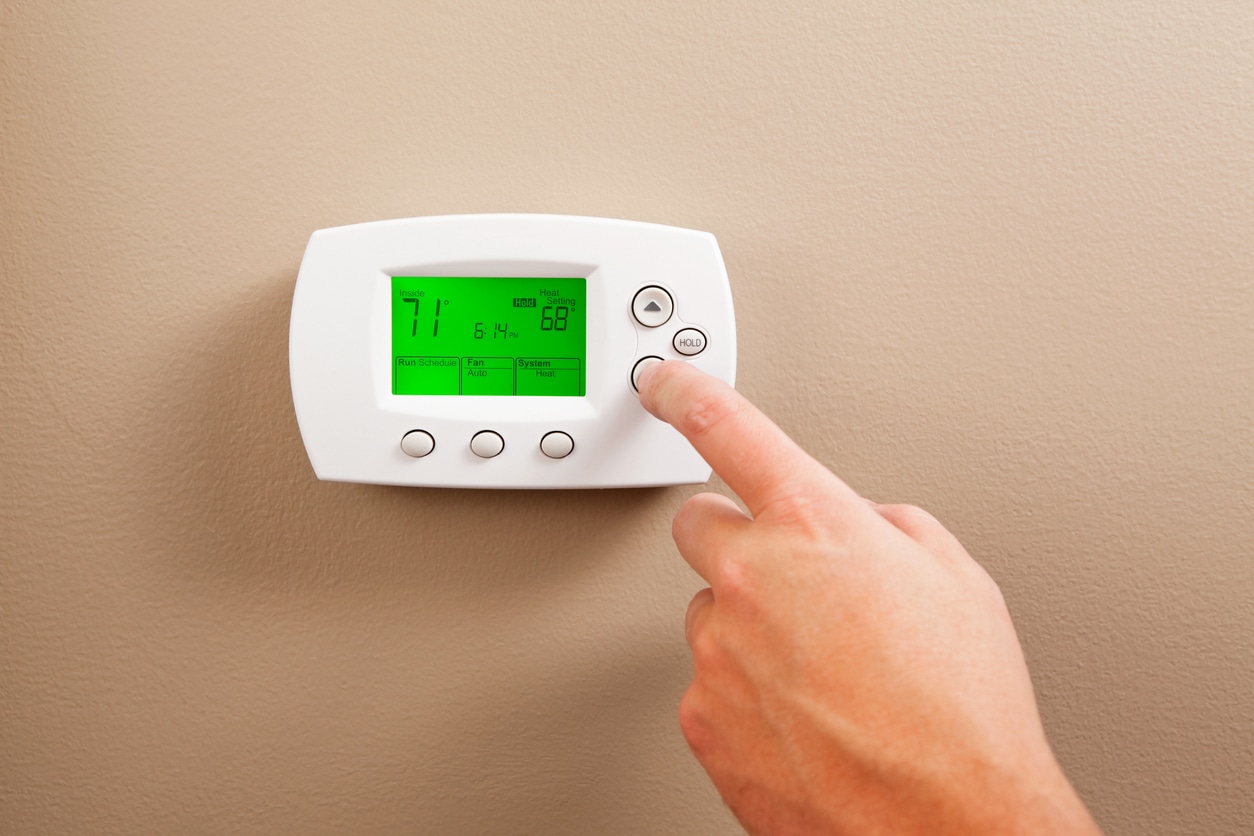 As a energy saving measure a male hand is turning down a digital programmable thermostat. The temperature reads 71 degrees, the adjusted temperature on the right is 68. Turning a furnace down while away or at night reduces electricity and gas consumption.