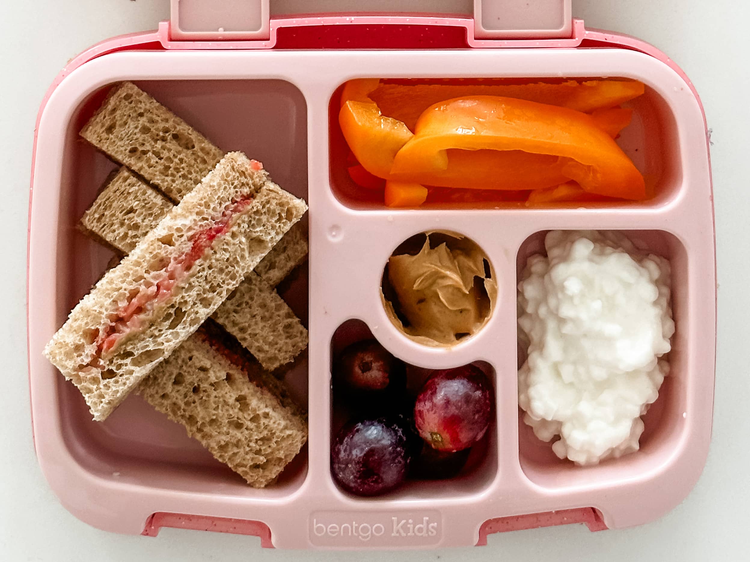 Kid's pink bento box with Sun Butter and Fruit Sticks with cottage cheese, bell peppers, grapes and peanut butter