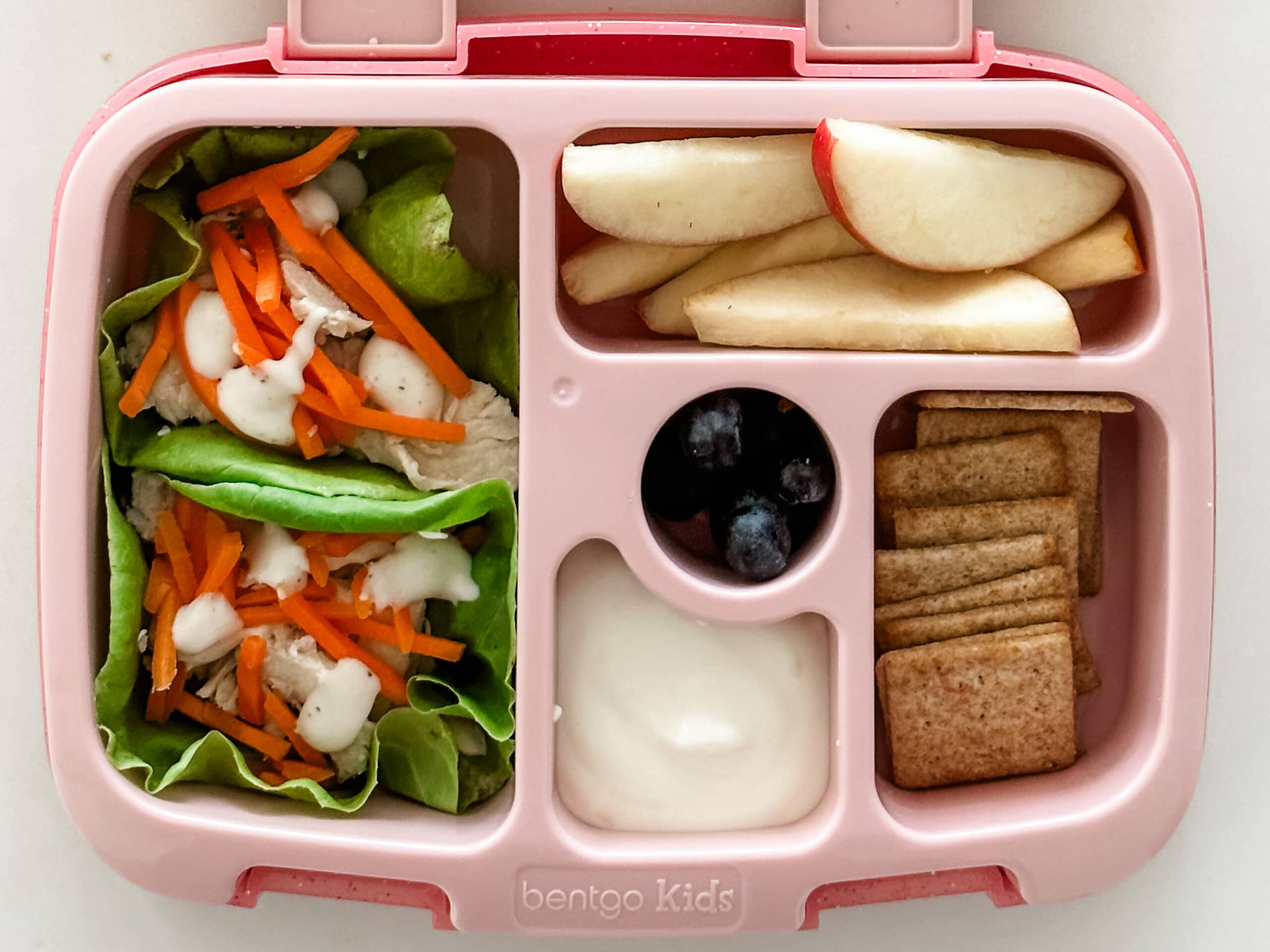 Kid's pink bento box with Lettuce Cups With Grilled Chicken, yogurt, crackers, apple slices, and blueberries