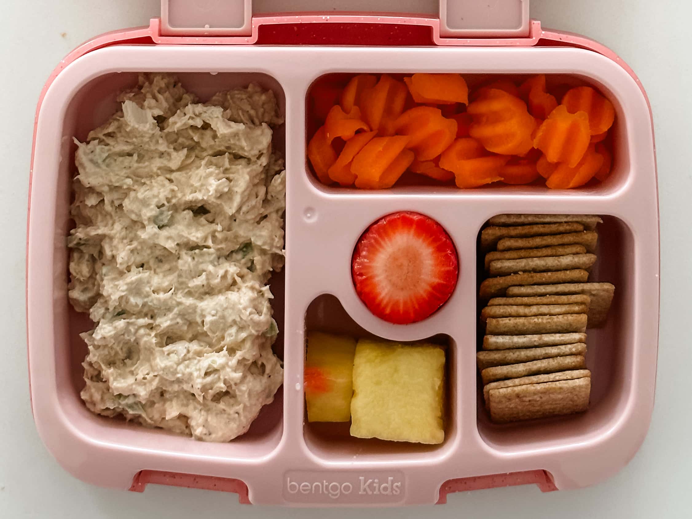 Kid's pink bento box with Chicken Salad, carrot chips, crackers, pineapple, and strawberry