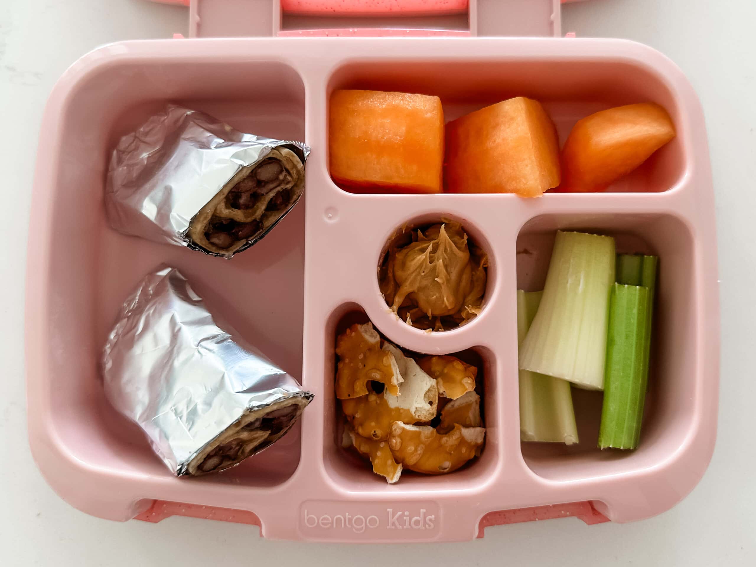 Kid's pink bento box with bean and cheese burrito and cantaloupe, celery, pretzels, and peanut butter