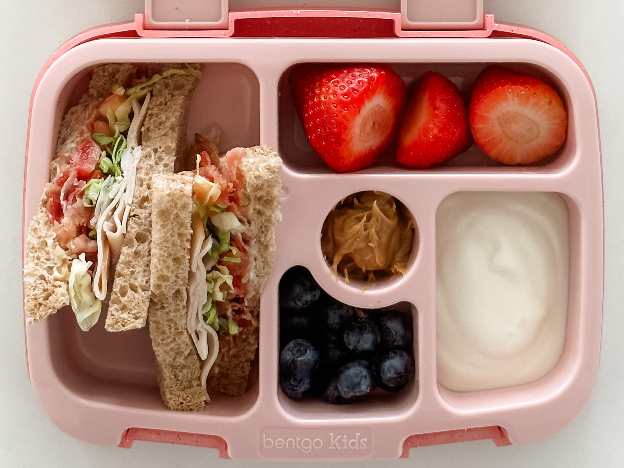 Kid's pink bento box with turkey club sandwich, strawberries, blueberries, yogurt, and peanut butter