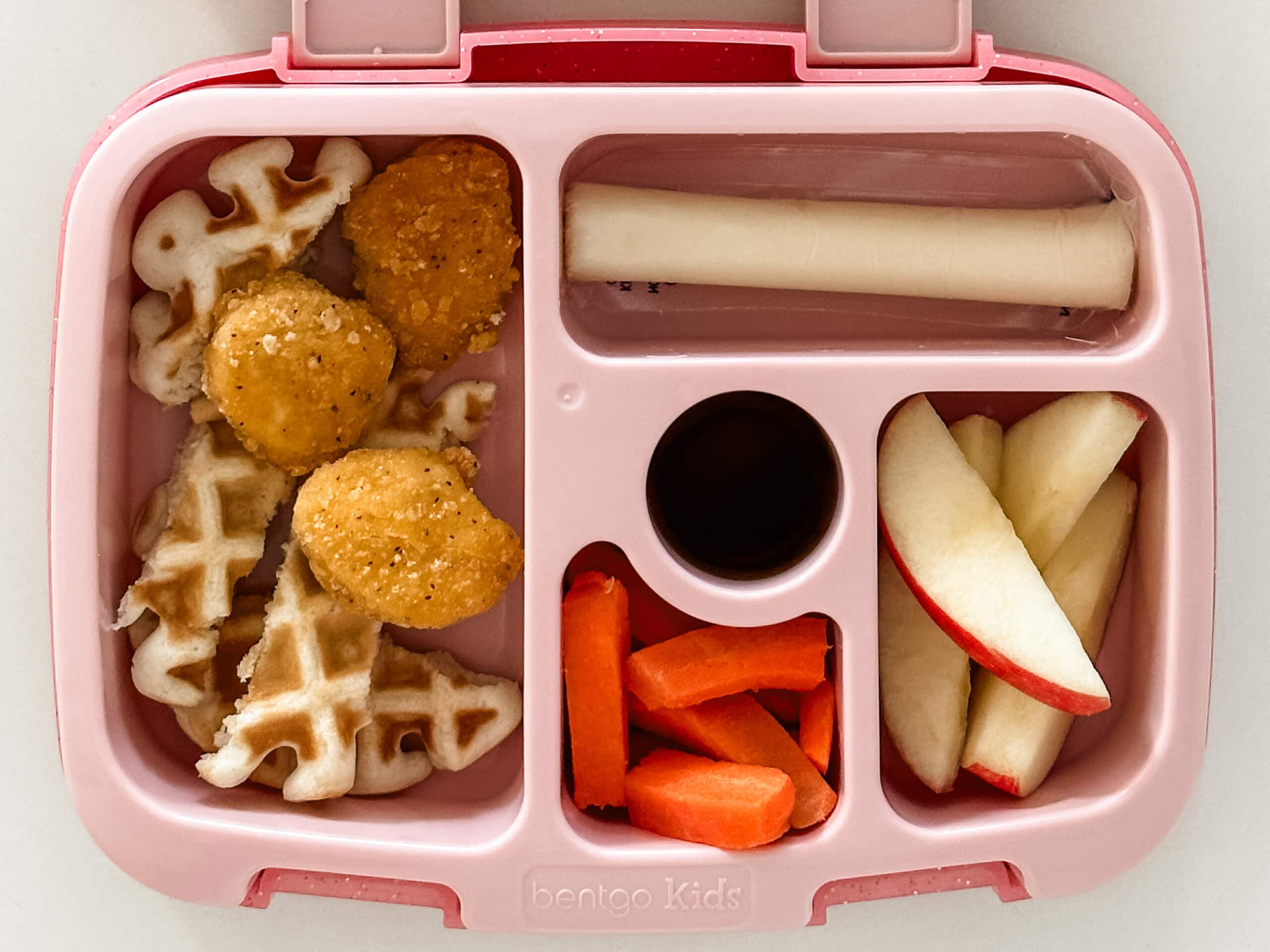 Kid's pink bento box with waffle strips, chicken nuggets, apple slices, carrots, cheese stick, and syrup