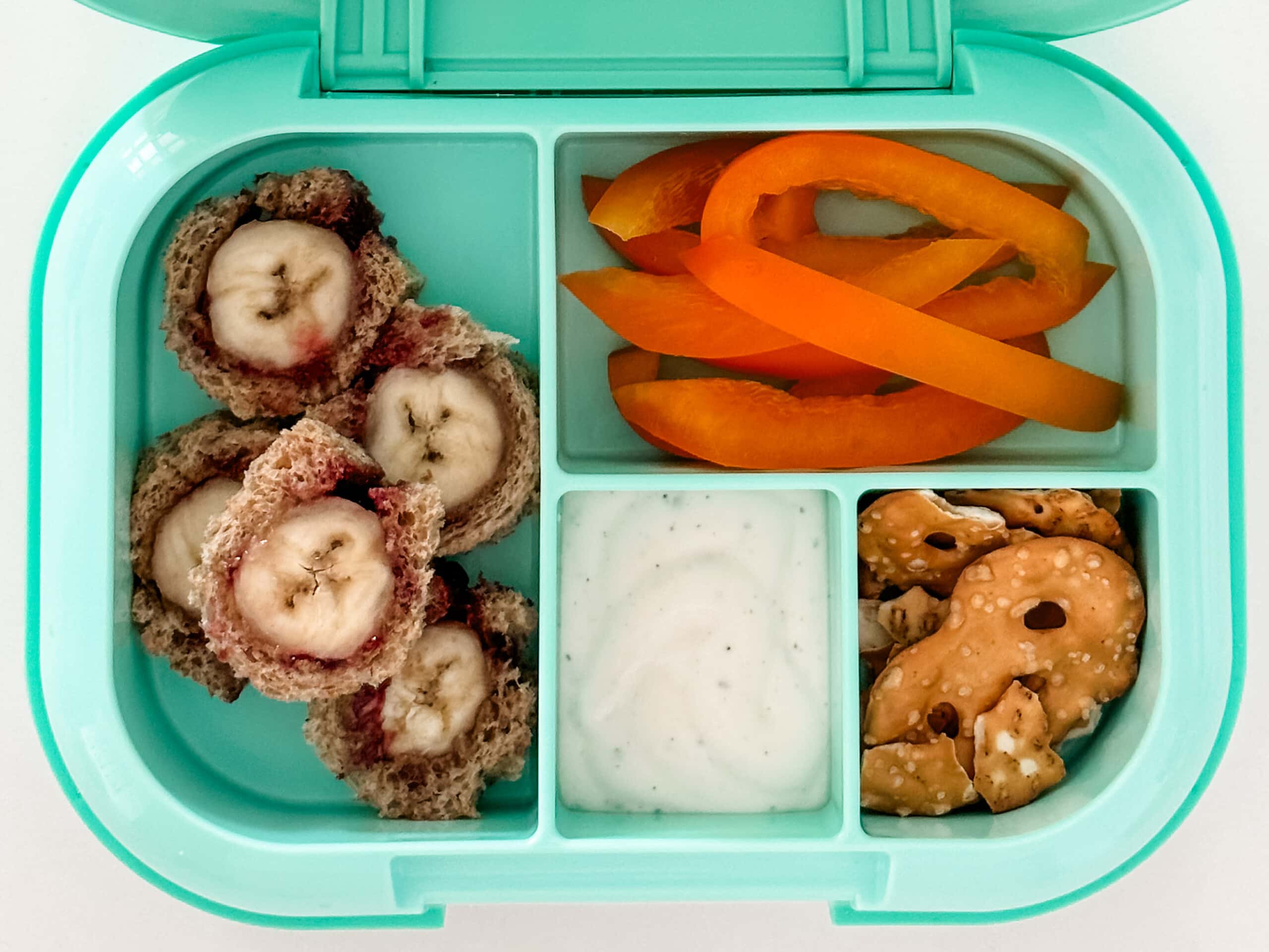 Mint green bento box for kids filled with banana roll ups, bell peppers, pretzels, and ranch dressing