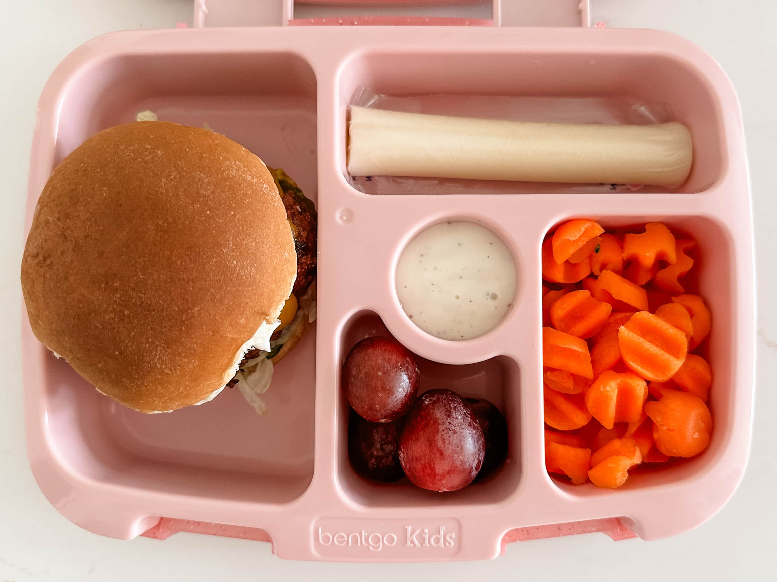 Kid's pink bento box with a slider, grapes, string cheese, carrot chips, and ranch dressing