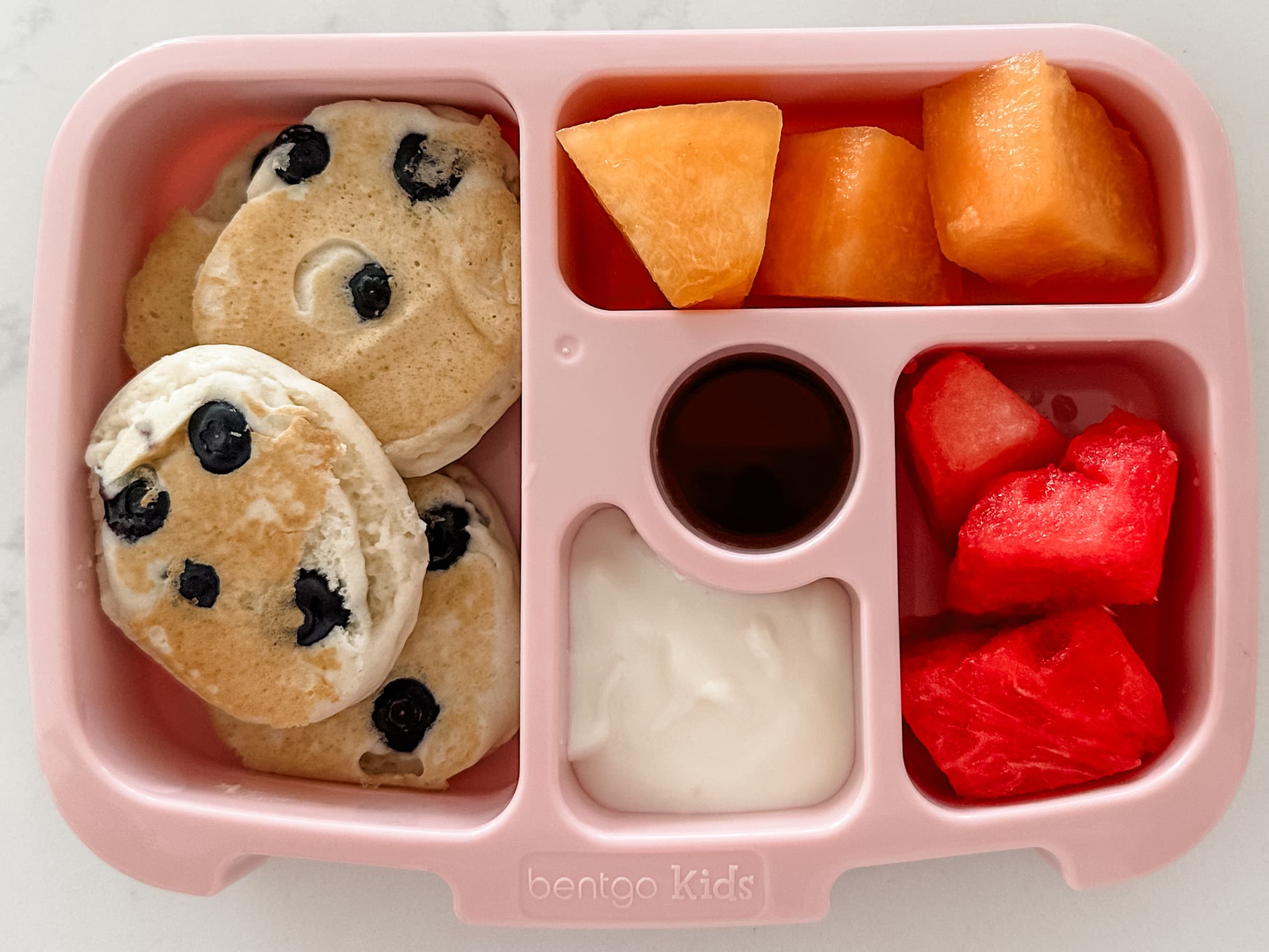 Kid's pink bento box with blueberry pancakes, cantaloupe, watermelon, yogurt, and syrup