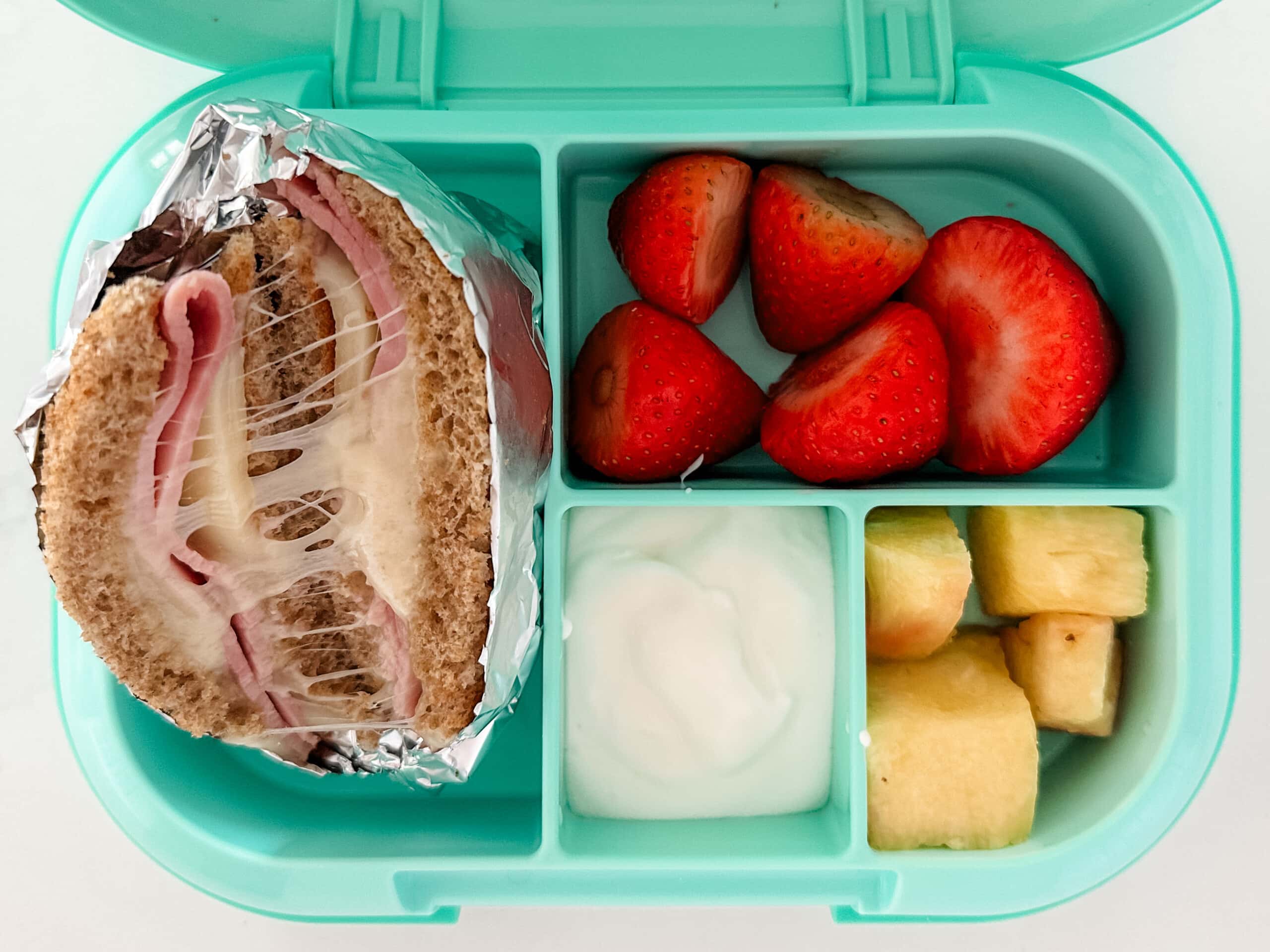 Mint green bento box for kids filled with hot ham and cheese sandwich wrapped in foil with strawberries, pineapples, and yogurt