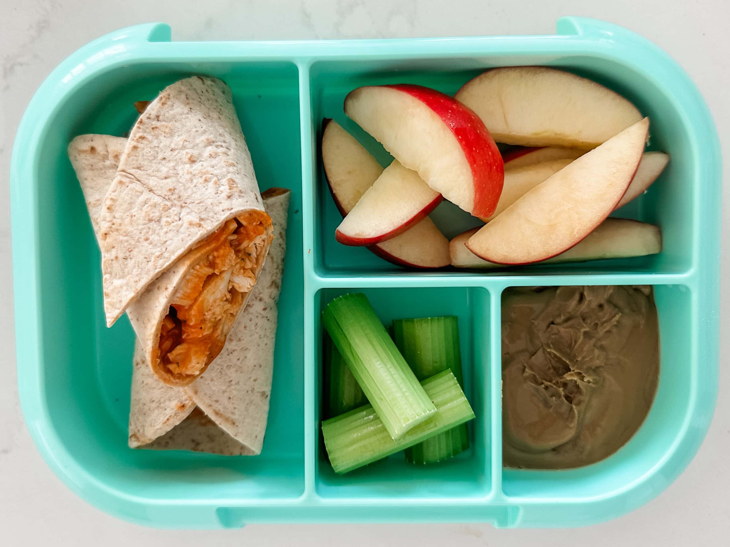 Mint green bento box for kids filled with a buffalo chicken wrap, apple slices, celery sticks, and sun butter