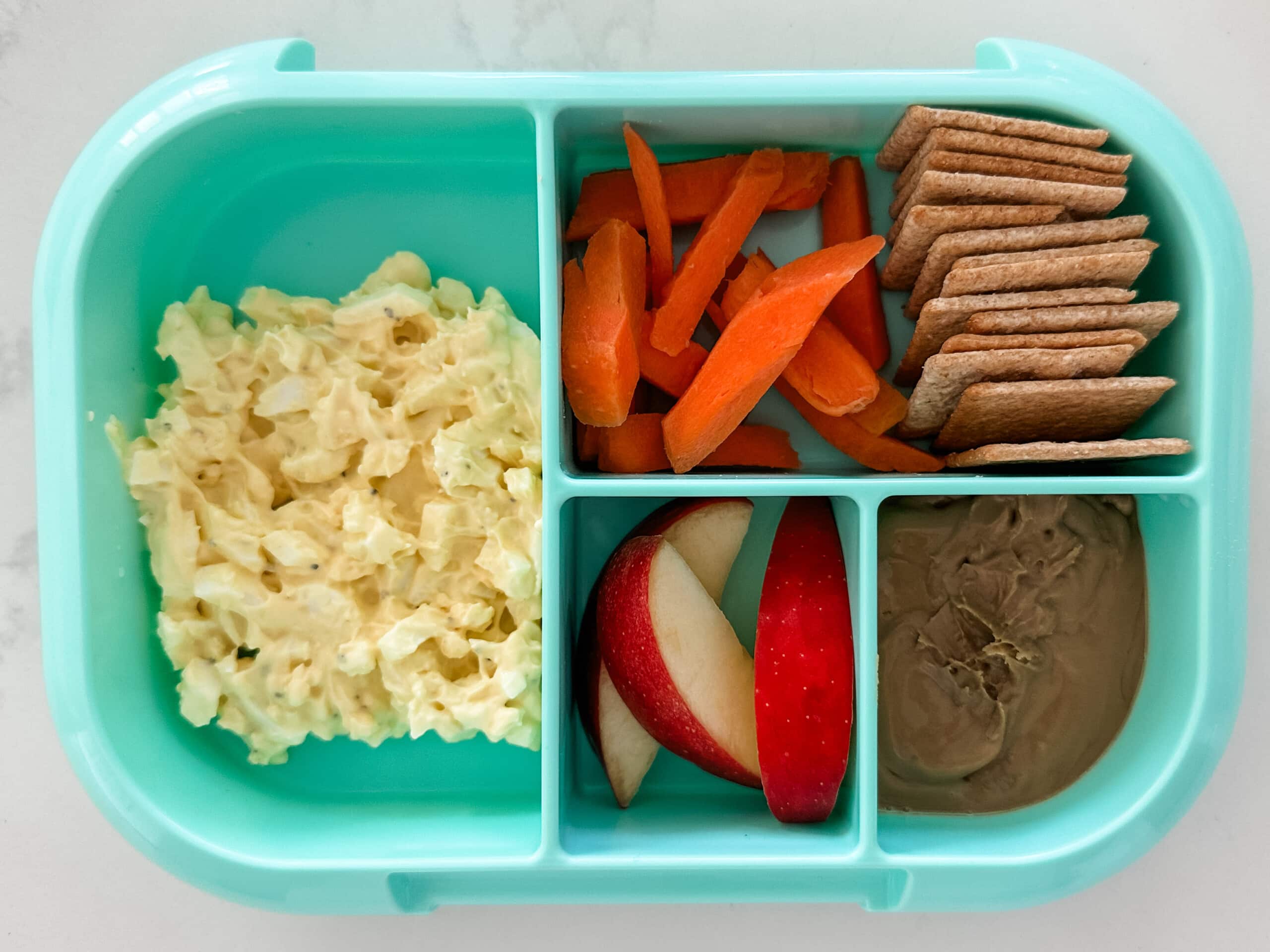 Mint green bento box for kids filled with egg salad, carrots, crackers, apple slices, and sun butter