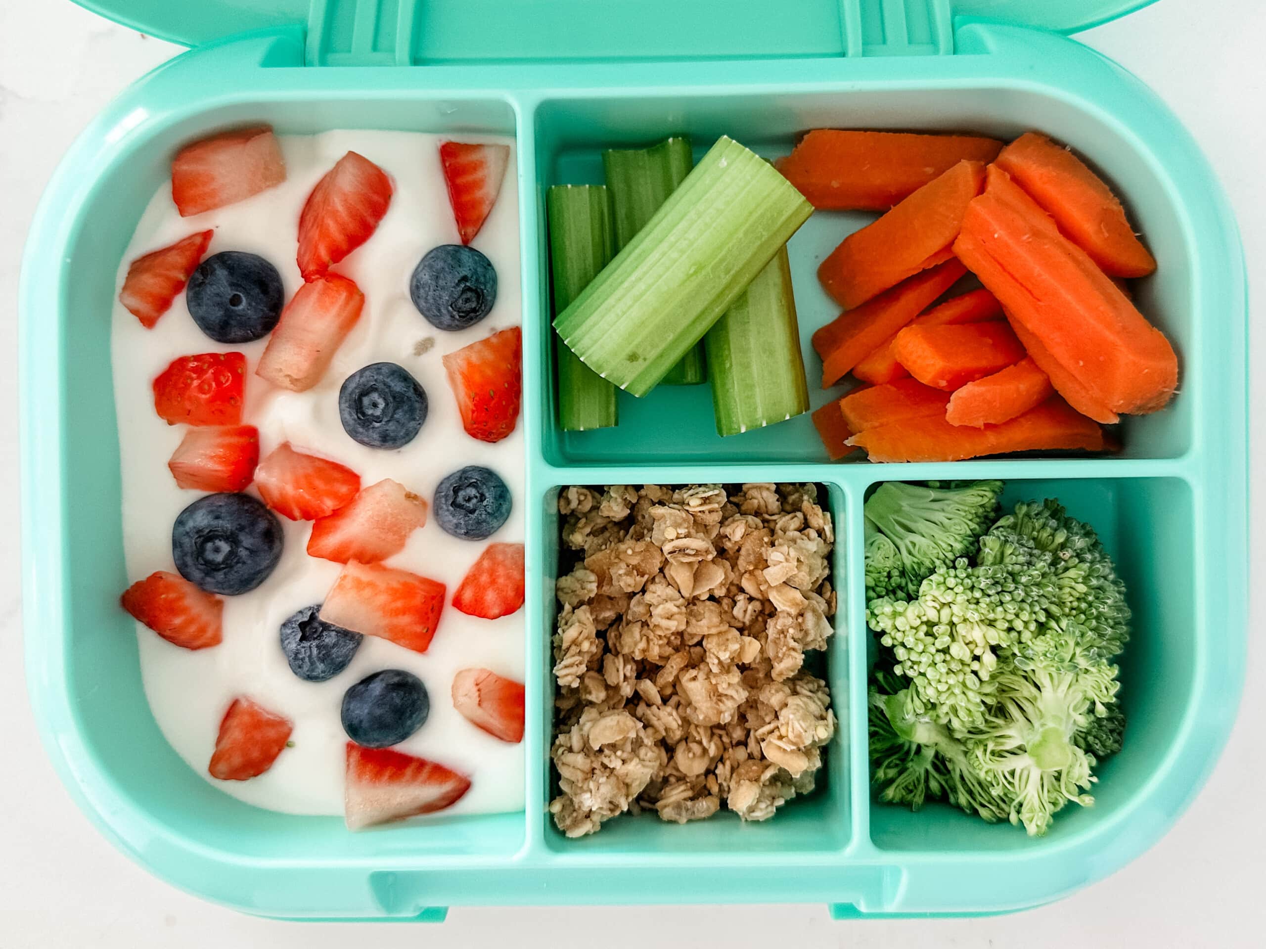 Mint green bento box for kids filled with greek yogurt and berries, granola, broccoli, celery sticks, and carrots