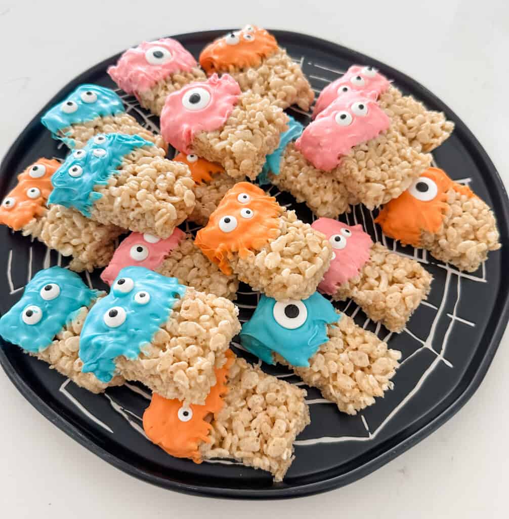 A black plate holds Monster Rice Krispie treats decorated with pink, blue, and orange icing. Each treat has candy eyes, giving them a monster-like appearance—perfect for creative Halloween snacks or fun Halloween dessert ideas.
