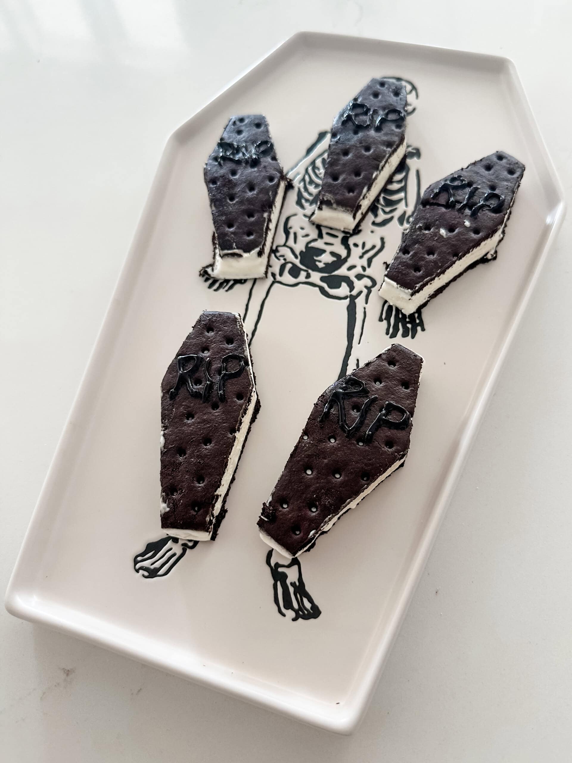 Five coffin-shaped ice cream sandwiches with RIP written on them are placed on a platter with a skeleton illustration, making the perfect Halloween desserts for a kids Halloween party.