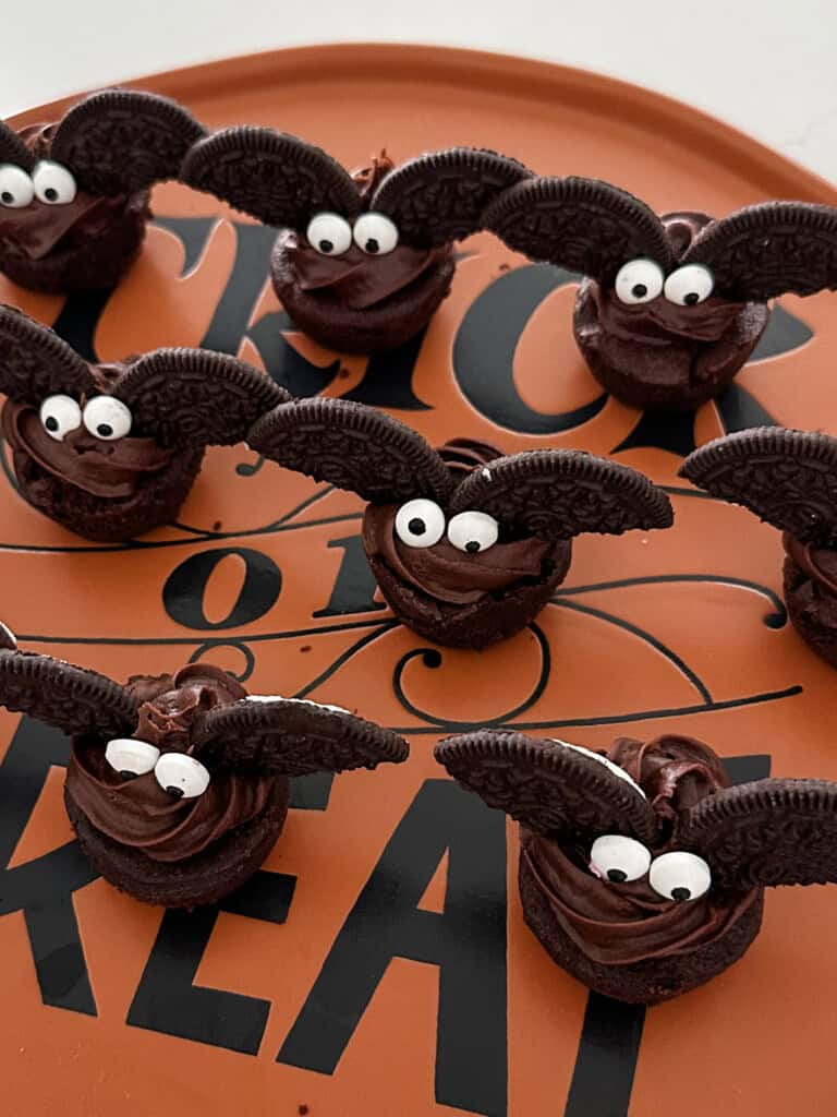 Chocolate mini brownies decorated to look like bats with Oreo wings and candy eyes are arranged on an orange Trick or Treat plate—a perfect addition to kids Halloween party snack ideas!