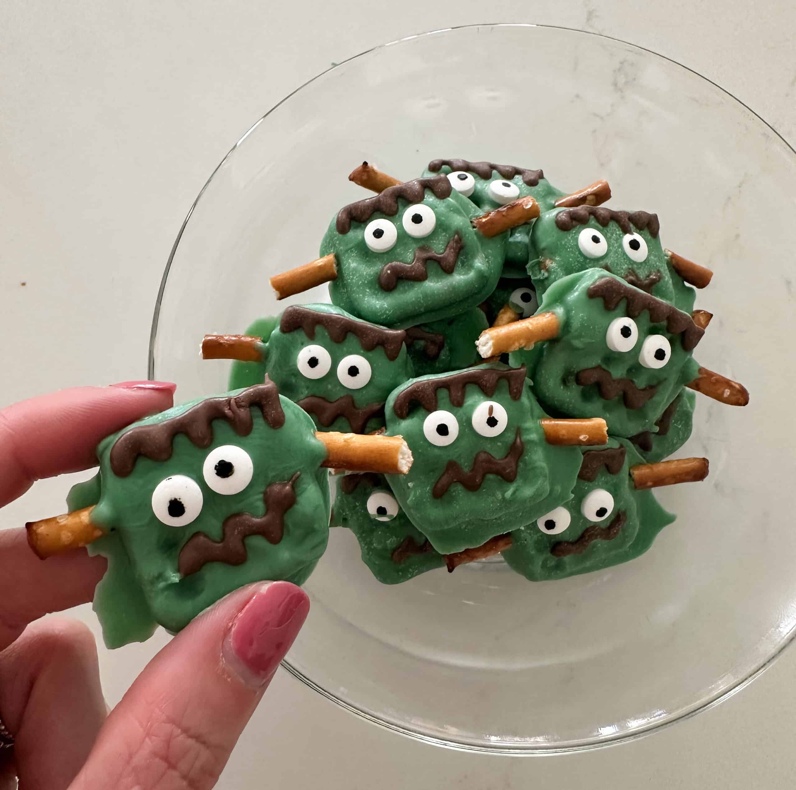 A bowl of spooky Frankenstein-themed Halloween treats, each with chocolate hair and eyes, and pretzel sticks resembling bolts. A hand is holding one treat. The delightful Halloween desserts rest on a clear glass dish against a light background, perfect for festive fun.