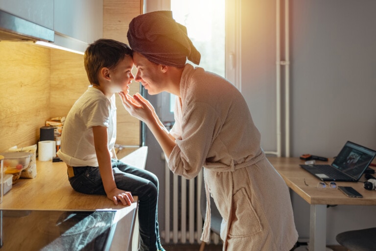 A young mom wearing a robe and a towel in her hair putting her head to her son's head. They are in the kitchen and looking into each other's eyes.