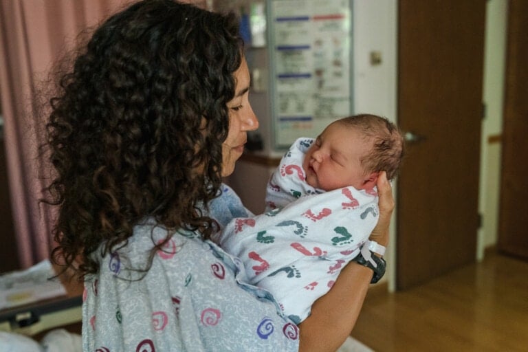 Profile view of a multiracial woman wearing a hospital gown standing in her hospital room while affectionately holding and lovingly admiring her newborn baby as he sleeps.