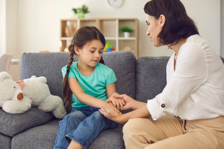 Young mother sitting on sofa with daughter, holding her hands and talking to her seriously at home with room interior at background. Solving problems in children education concept