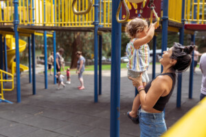 A young Latin American woman helps a two year old girl use the jungle gym at the playground on a warm sunny afternoon.