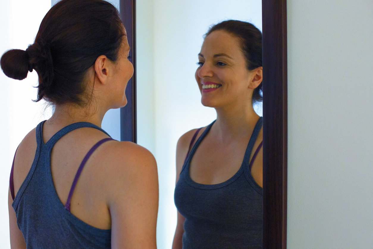 Woman practicing a smile looking herself in the mirror. Self-talk psychology method by Louise Hay.