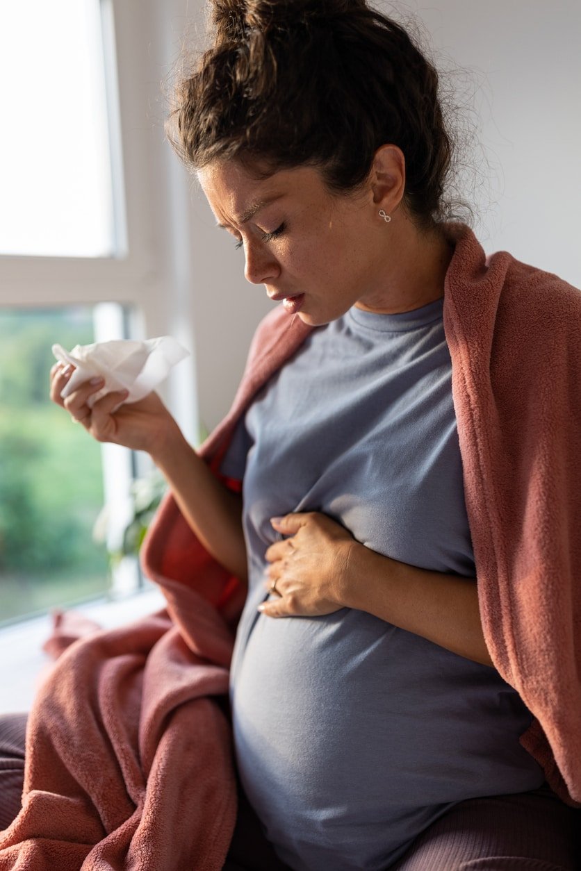Pregnant woman covered with blanket sitting on bed and sneezing into tissue.