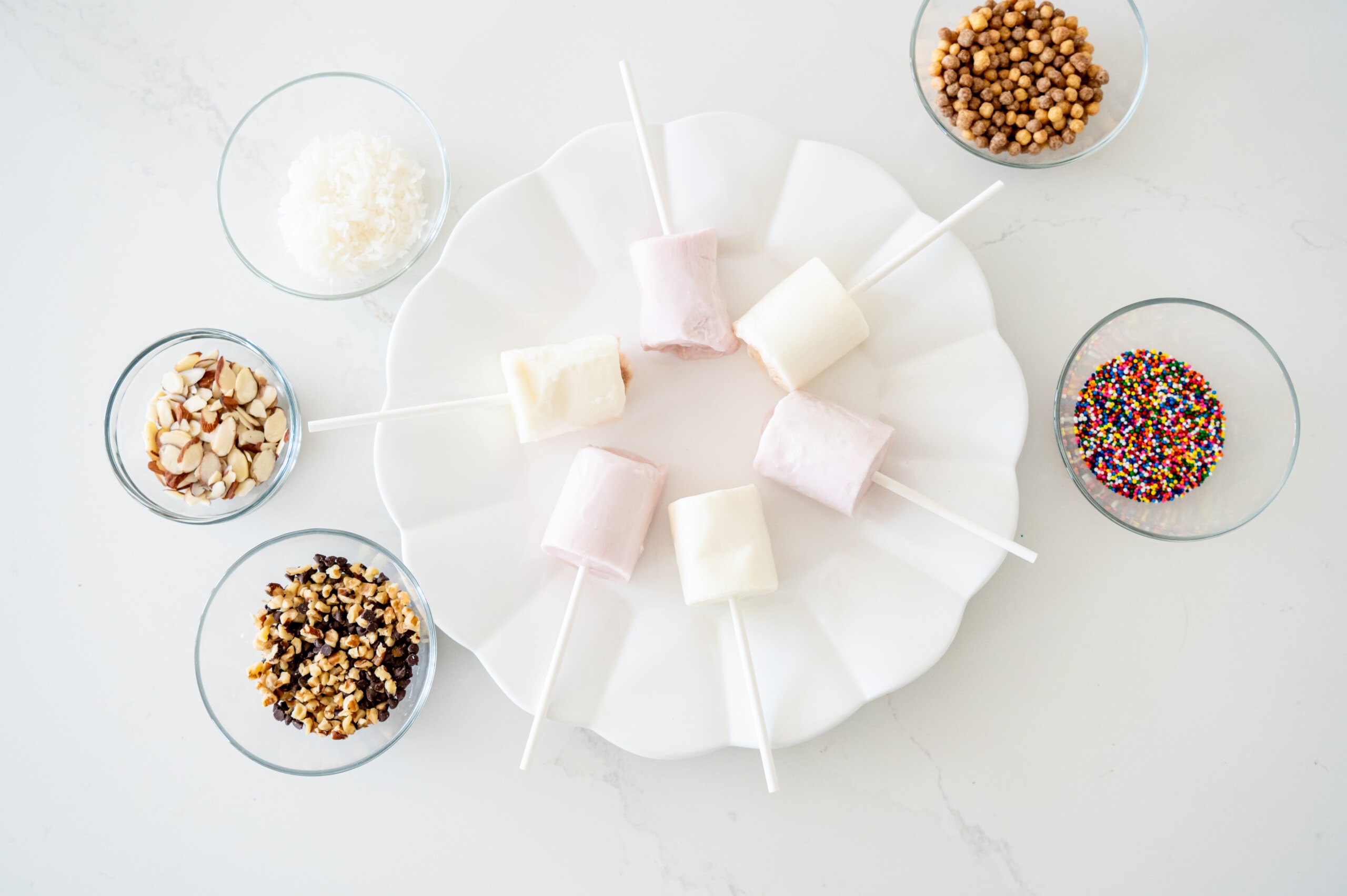 A white plate with yogurt pops and small bowls around with different topping choices.
