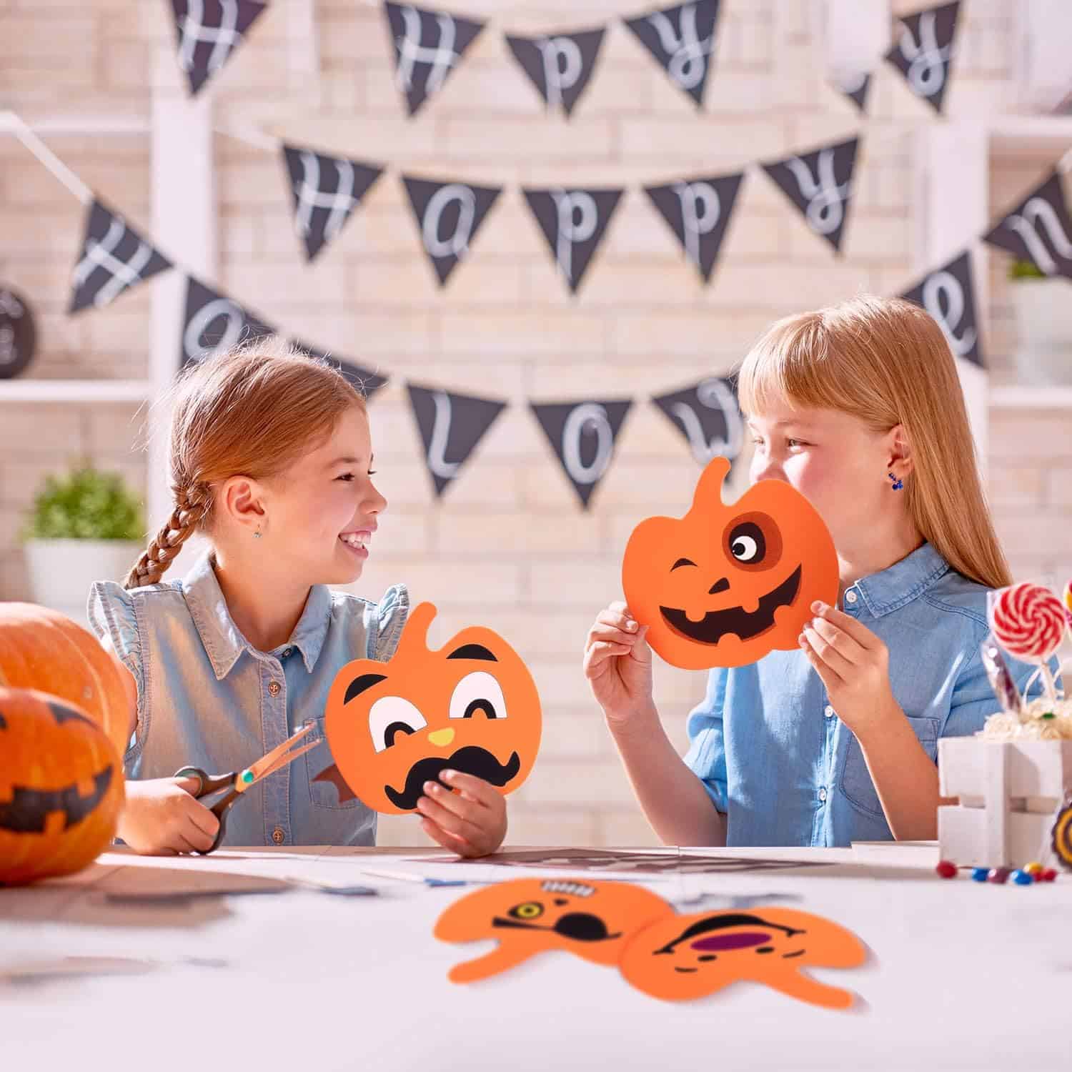Two girls are sitting at a table, smiling and holding pumpkin-shaped Halloween crafts. Both are dressed in blue shirts. Various craft supplies and Halloween decorations, perfect for Halloween activities for kids, are on the table with a Happy Halloween banner hung in the background.