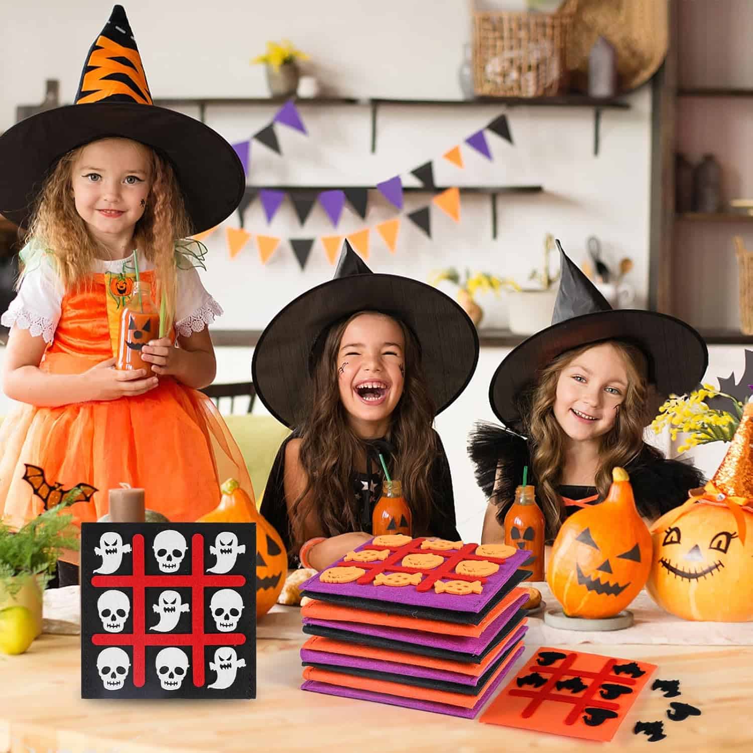 Three children dressed in witch costumes smile and laugh at a decorated table. The table features Halloween-themed items, including painted pumpkins and tic-tac-toe games with skull and bat designs—perfect Halloween activities for kids. Orange and black banners hang in the background.