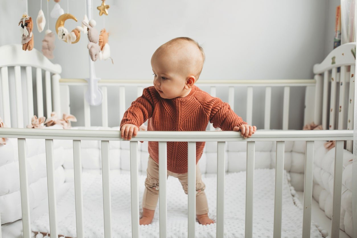 Beautiful little girl standing up in a crib in a cozy light room in a sweater of fashionable shades. learns to stand at the support. cheerful little girl standing in the crib at home