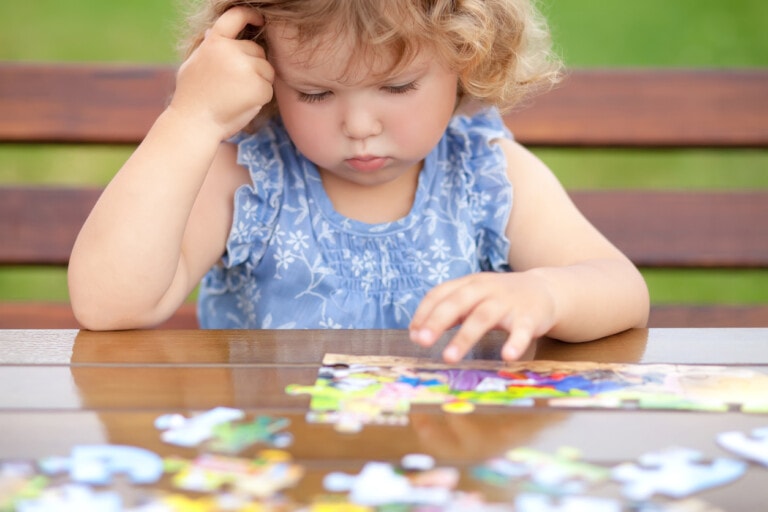 Blonde unhappy toddler girl, solving puzzle on a table, hard difficult task. Early education and development.
