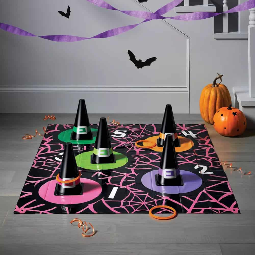 A Halloween-themed ring toss game setup in a room. Five black witch hats with colored bases are placed on a spiderweb mat. Rings and a pumpkin carved with stars are on the floor. Purple streamers and black bat decorations hang in the background, perfect for Halloween activities for kids.