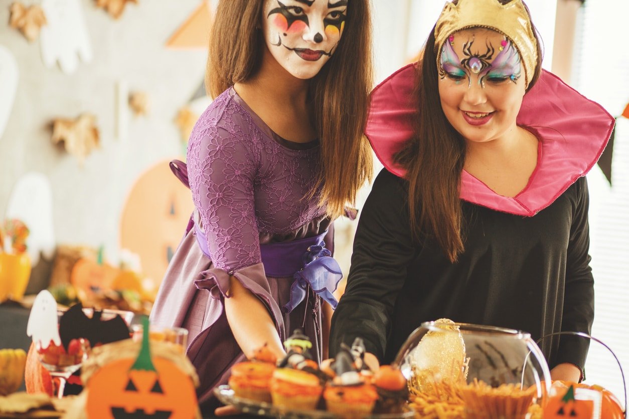 Two girls in Halloween costumes are standing at the buffet full with candies, snacks, and cupcakes.