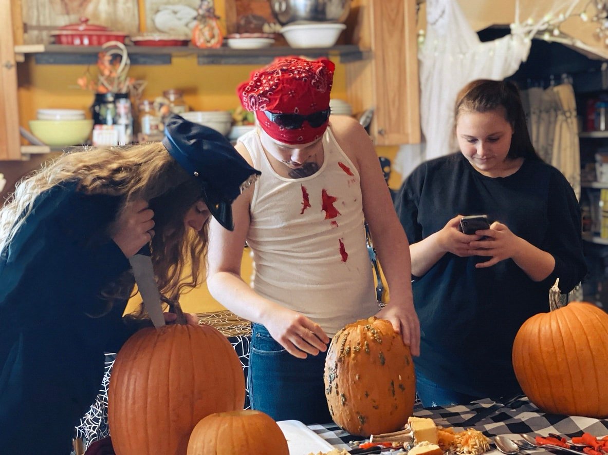 Group of teens(siblings and a friend) at home, in the kitchen at a Halloween pumpkin carving party. The teens are wearing costumes and carving pumpkins, all busy doing something different. One teen girl is checking her phone.
