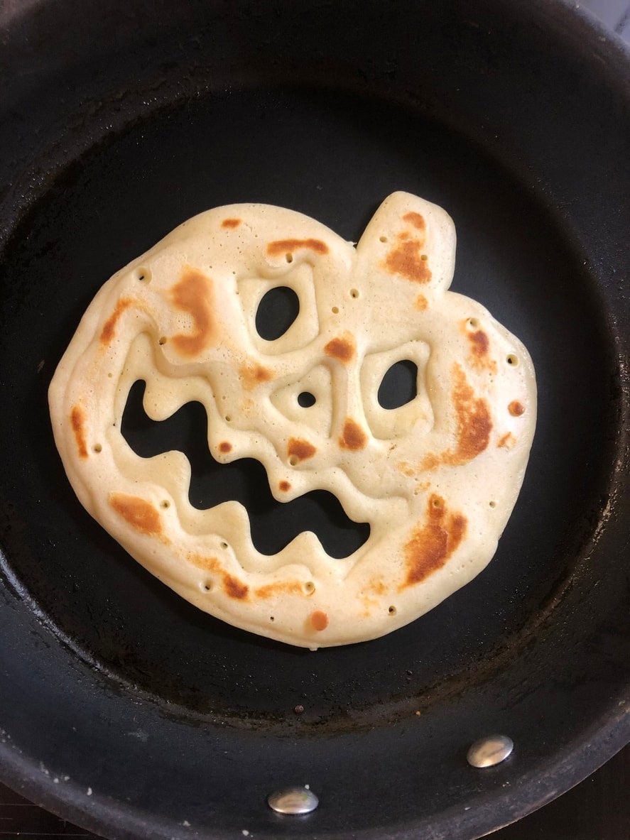 This picture is of a non-stick frying pan with a pancake shaped like a Jack-o'-lantern pumpkin.