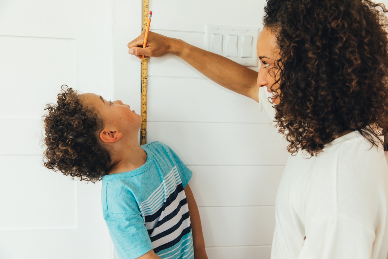 A mom measures her son against a ruler and wall to see how tall he is inside their home. The boy is growing fast at 4 years old and is excited to grow and learn with his mom teaching him.
