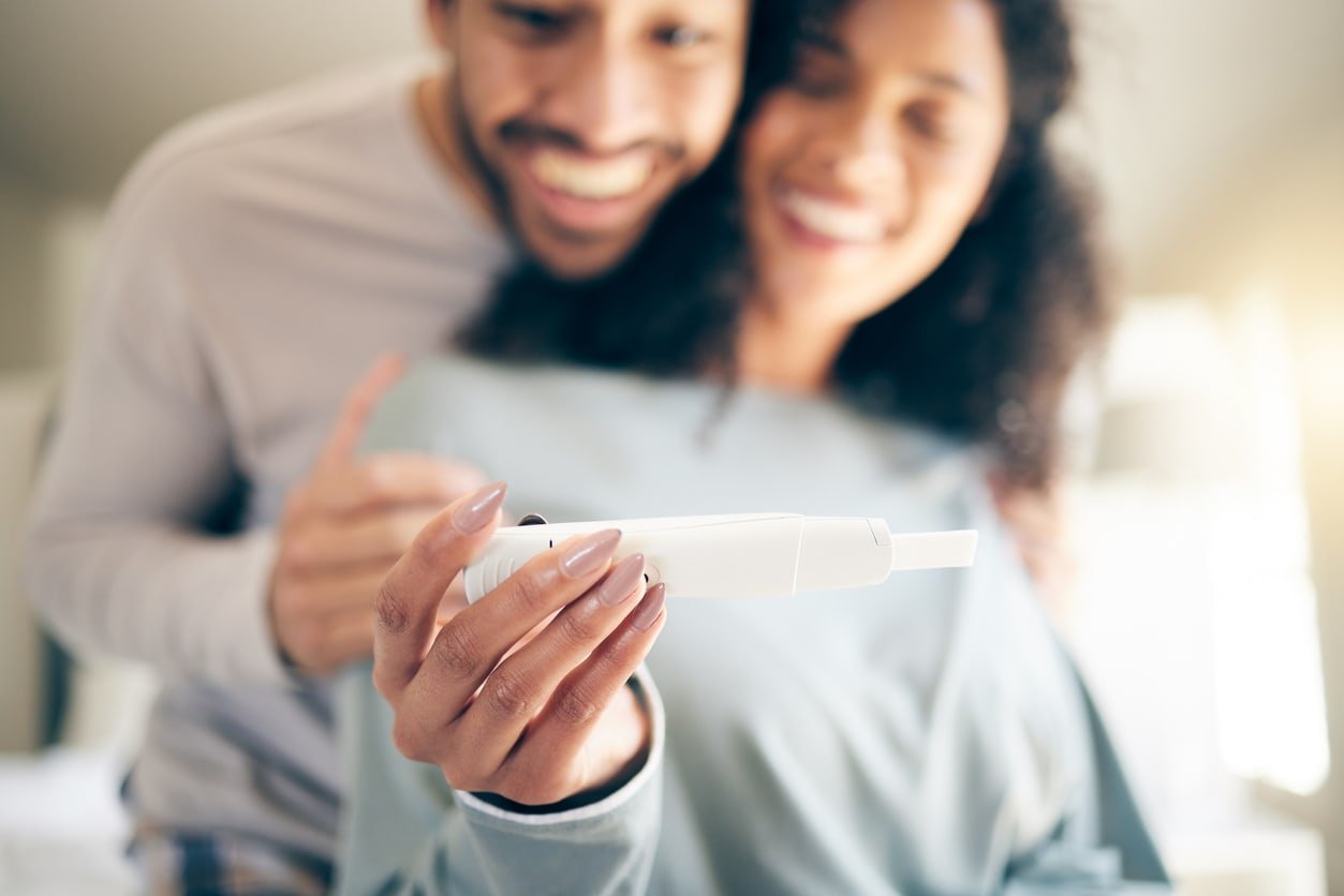 Pregnancy test, hands of woman and happy couple, smile and baby announcement on blurred background in home. Pregnant, person and new mother, fertility and support for positive results and ivf success