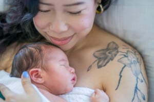 A new Mother holds her infant to her chest as they bond skin-to-skin in the comfort of their own home. The baby is wearing a diaper and both have their eyes closed as they rest peacefully.