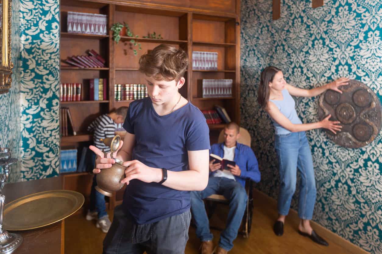 Four people are in a room with high bookshelves and patterned wallpaper, engaging in an activity. One person is examining an old kettle, another is sitting and using a device, a third is inspecting a wall fixture, and the fourth is looking at shelves—perfect inspiration for Halloween party ideas.