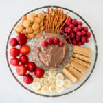 A glass bowl filled with a chocolate peanut butter dip with some raspberries on top. It's surrounded by fruits, cookies, and pretzels on a large dish.