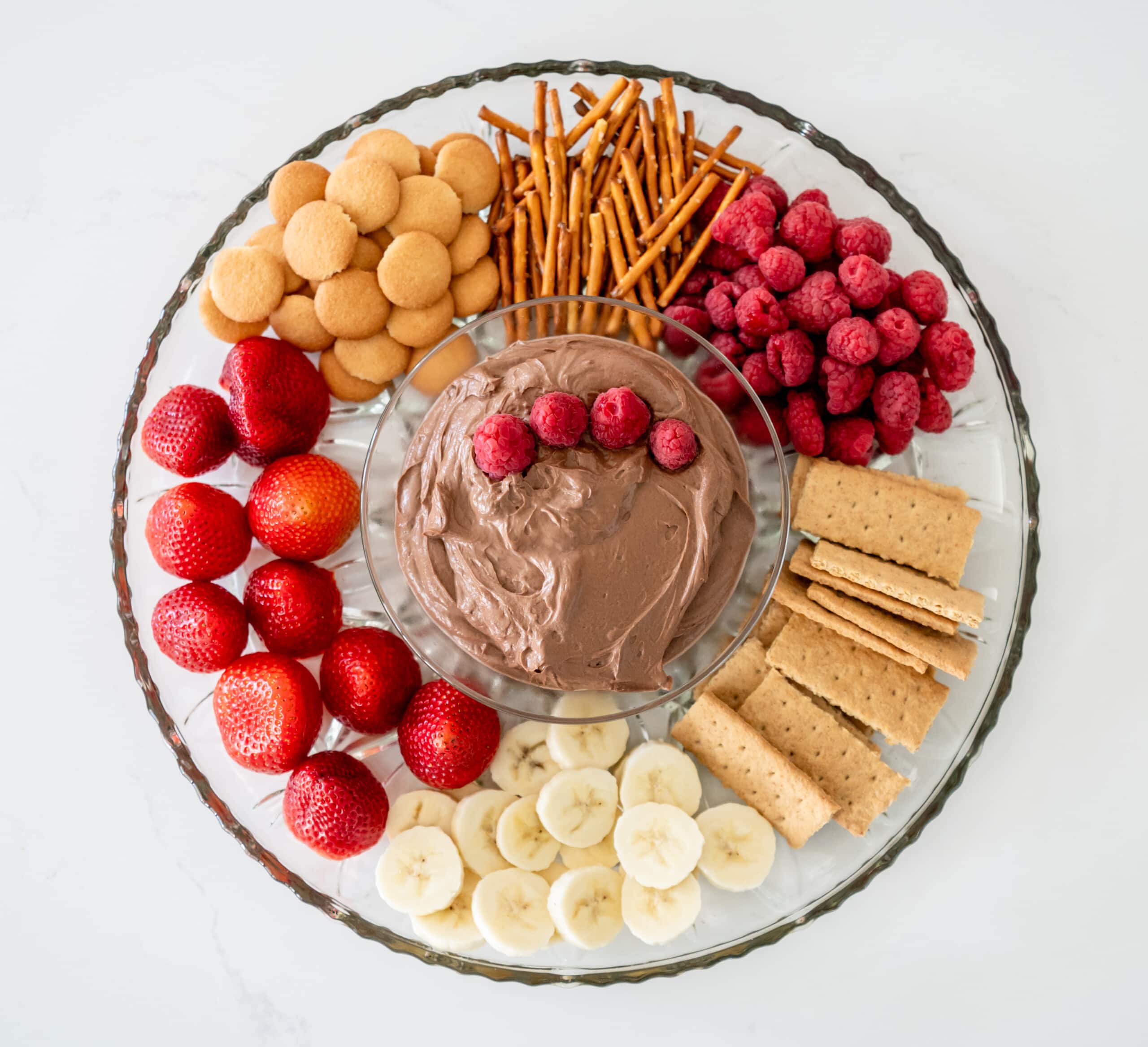 A glass bowl filled with a chocolate peanut butter dip with some raspberries on top. It's surrounded by fruits, cookies, and pretzels on a large dish.