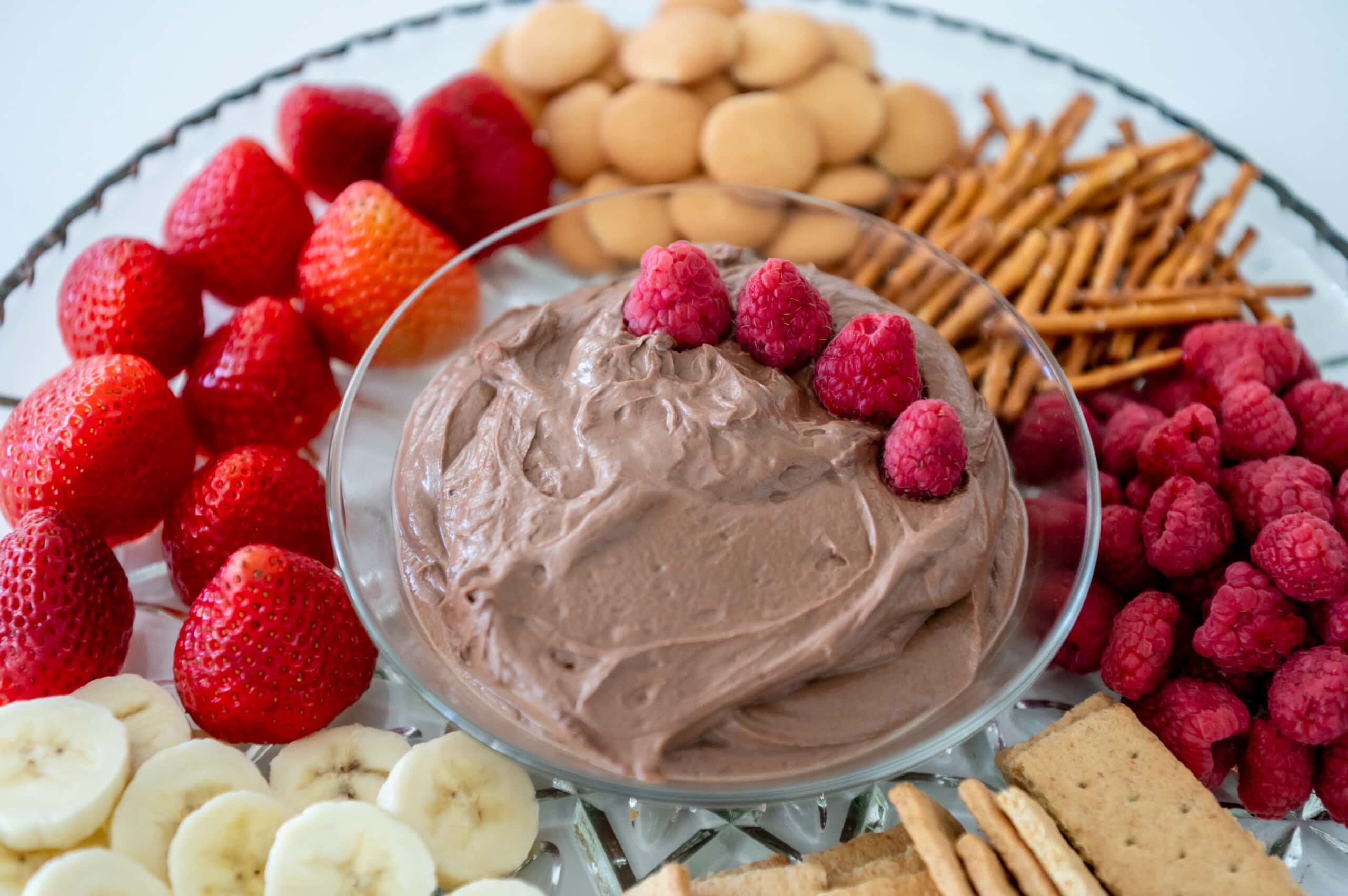 Close up of a glass bowl filled with a chocolate peanut butter dip with some raspberries on top. It's surrounded by fruits, cookies, and pretzels on a large dish.