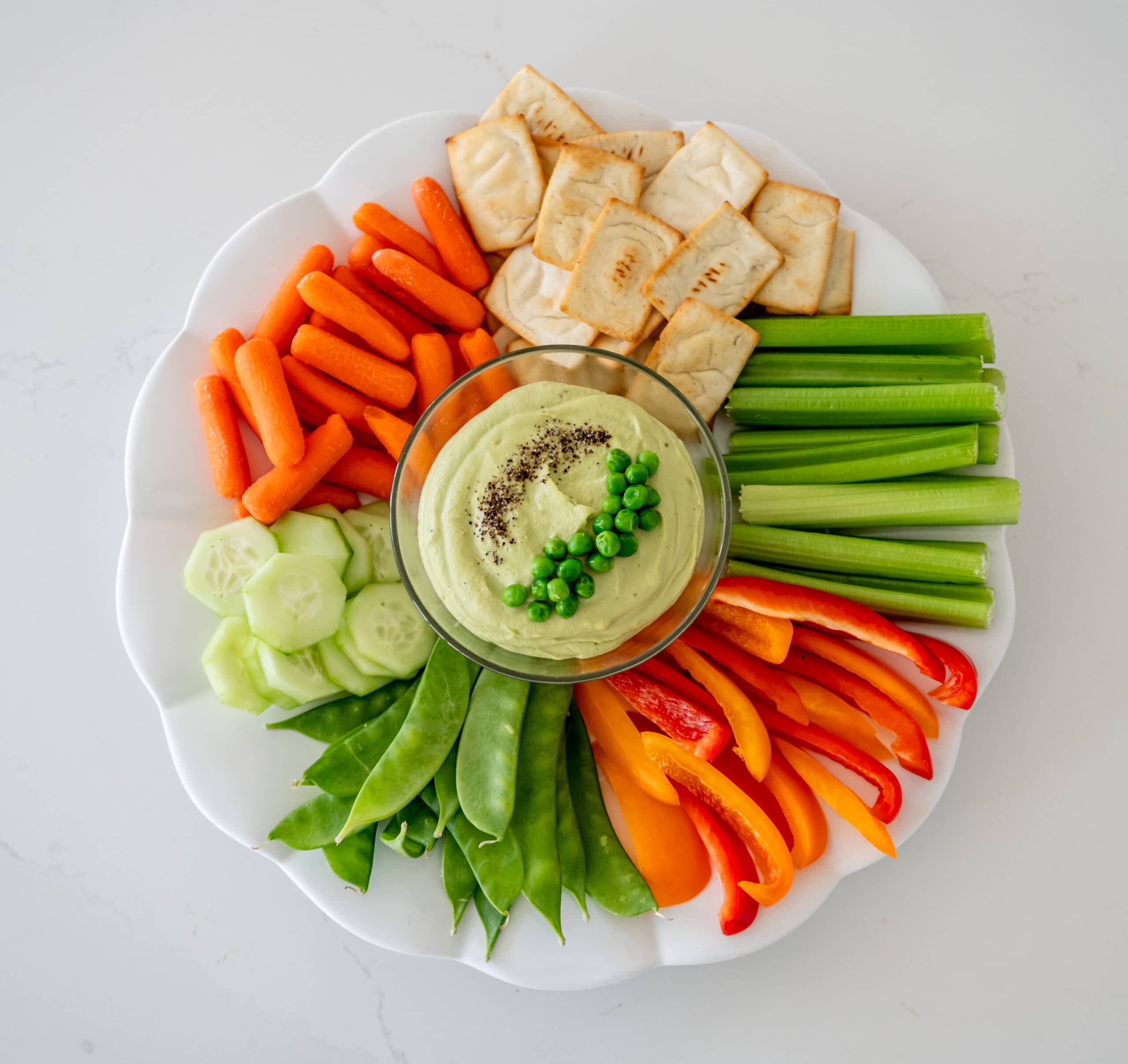Green pea hummus in the center of a white plate surrounded by veggies and pita chips.