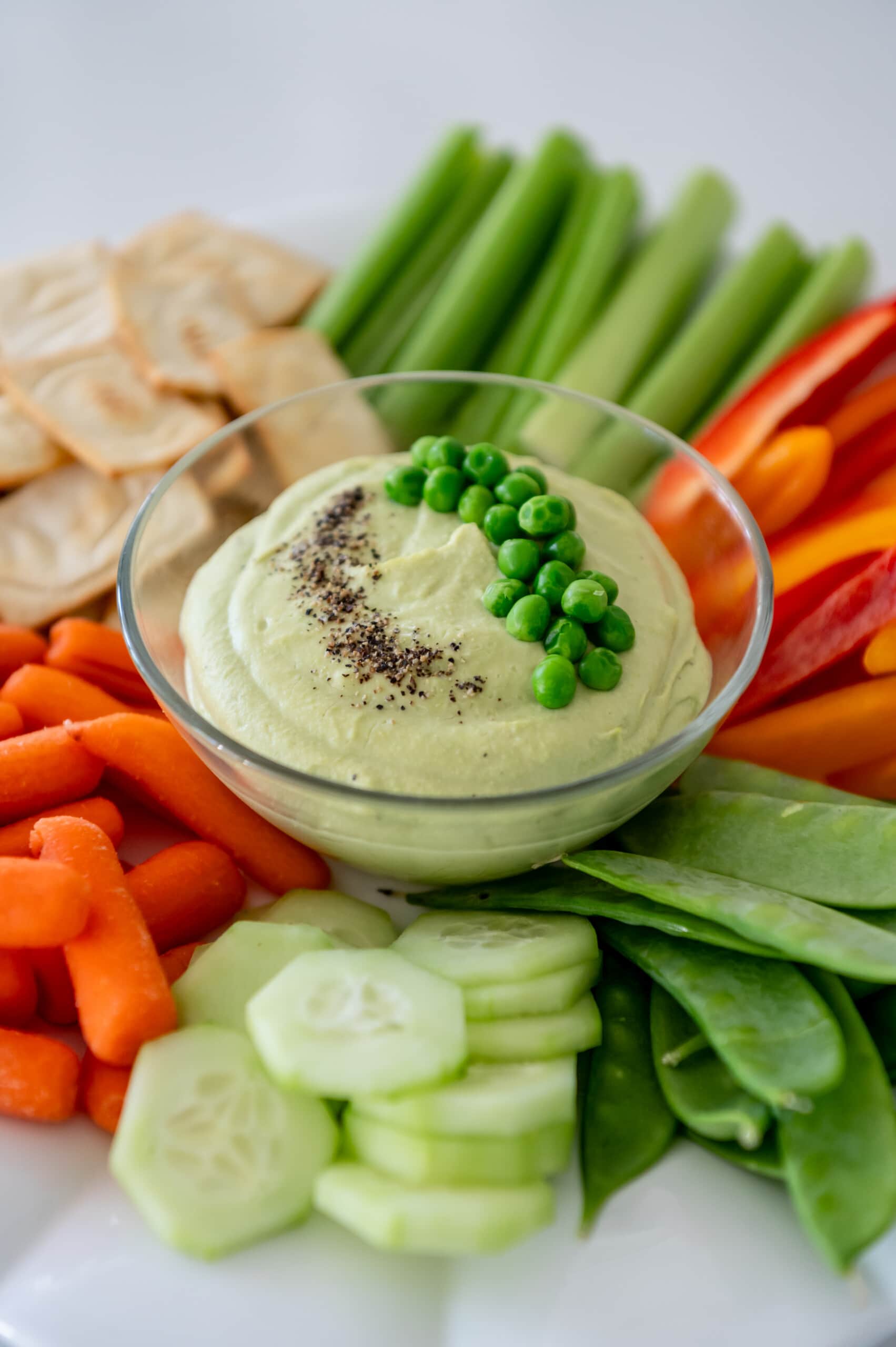 Close up of a glass dish that has green pea hummus inside. It is surrounded by veggies and pita chips.