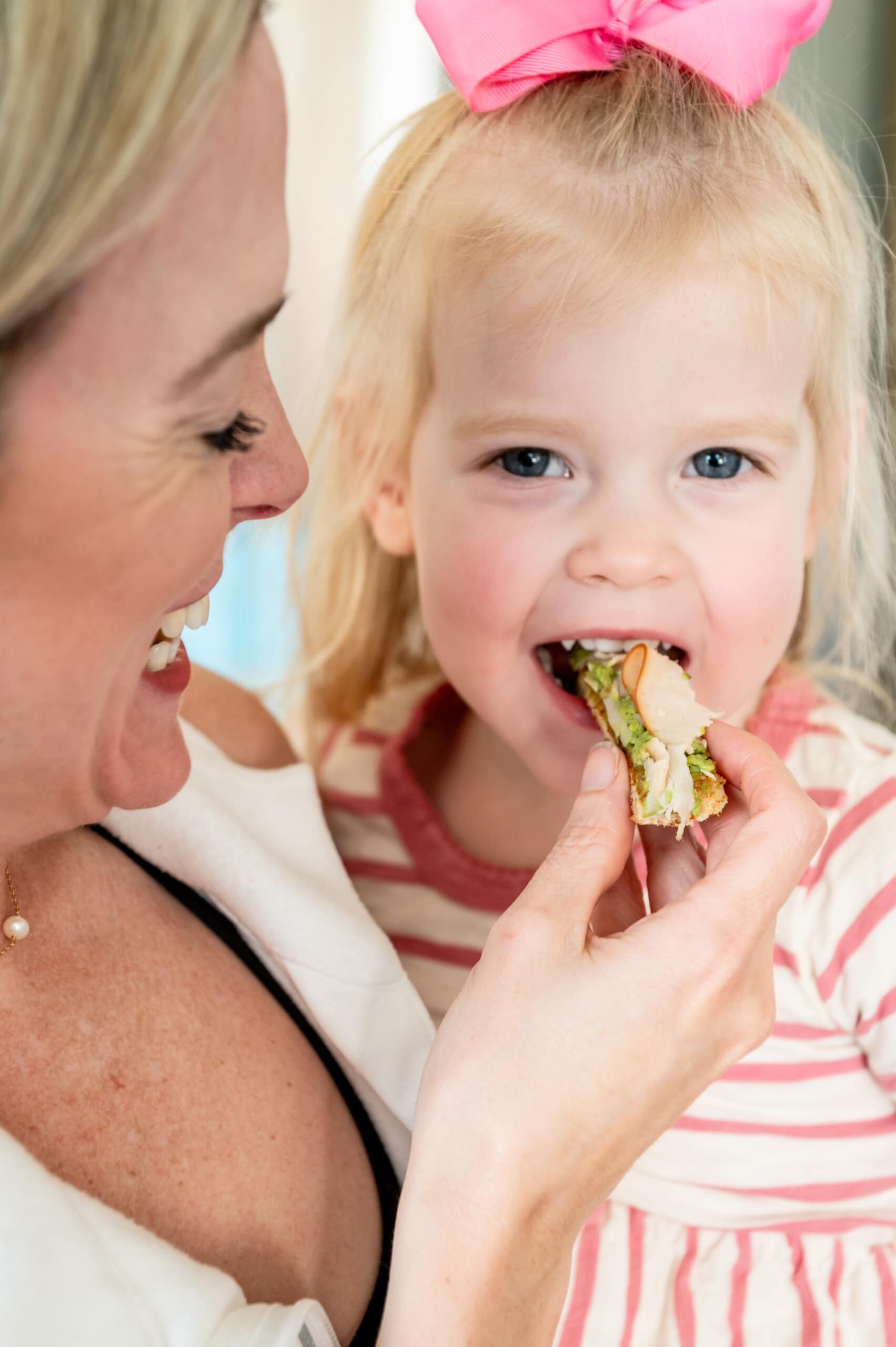 Toddler girl being held by her mom eating a bite of her avocado turkey toast
