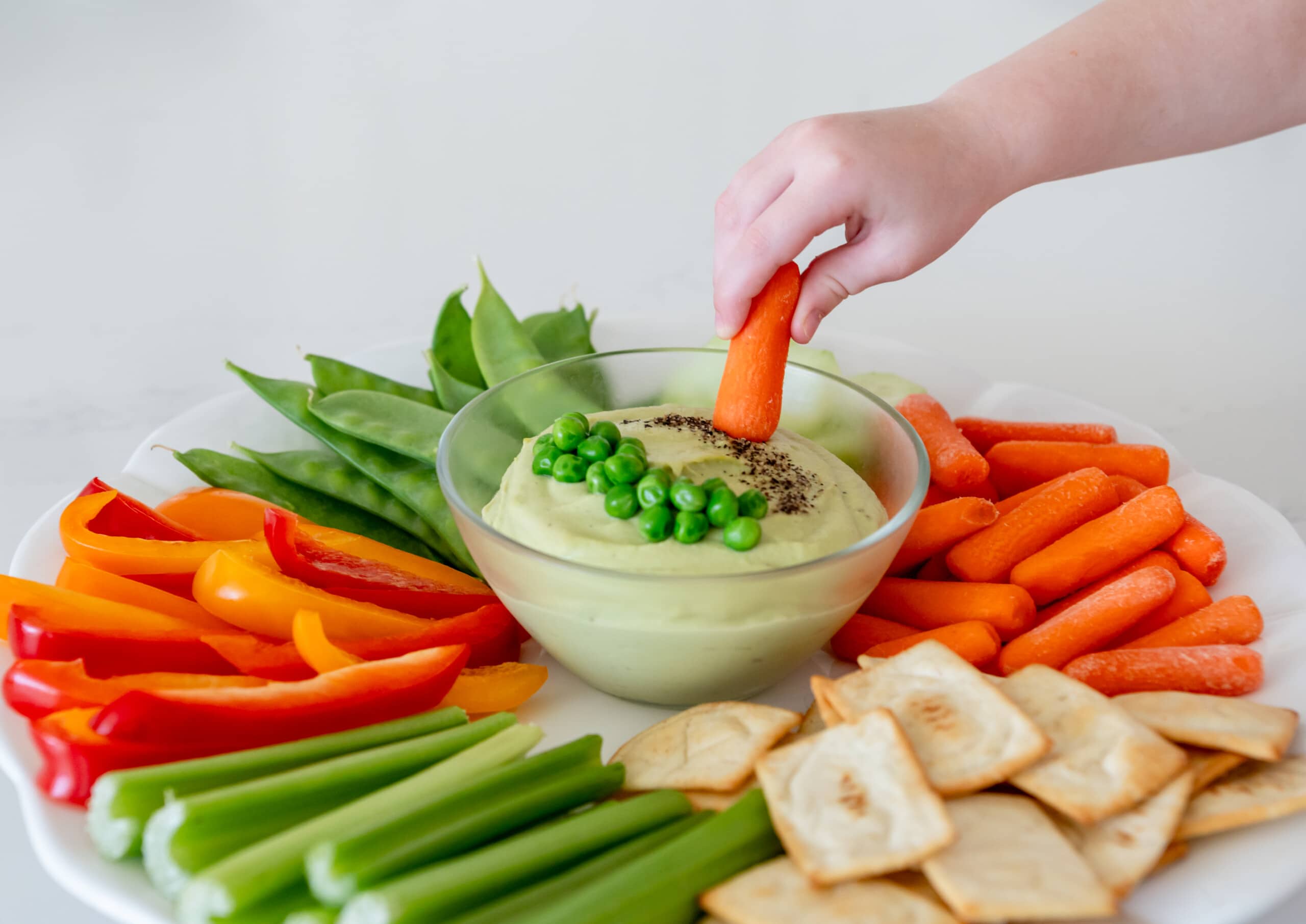 Little hand dipping a carrot into a bowl of green pea hummus.